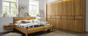Schlafzimmer mit Massivholzbett, schwebendem Nachttisch und großem Kleiderschrank aus Holz vor zwei Fenstern.